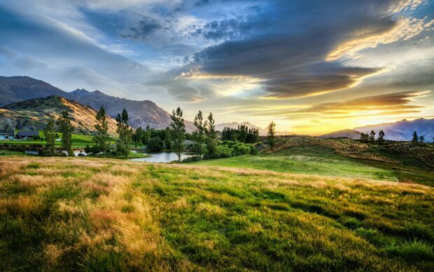 A scenic view of New Zealand with green fields and mountains under a dramatic sky at sunset