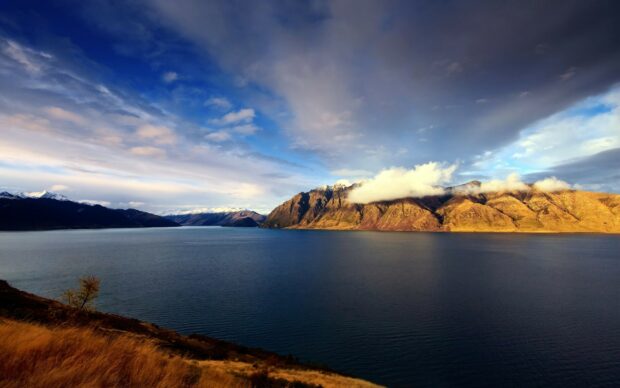 A scenic view of New Zealand mountains with clouds over a calm lake and golden grass in the foreground