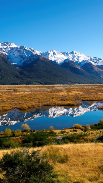 Snow capped mountain range in New Zealand reflected in a calm river surrounded by autumn vegetation