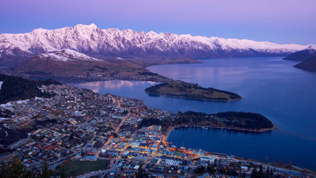 A panoramic view of New Zealand town with snowy mountains and lake under clear sky