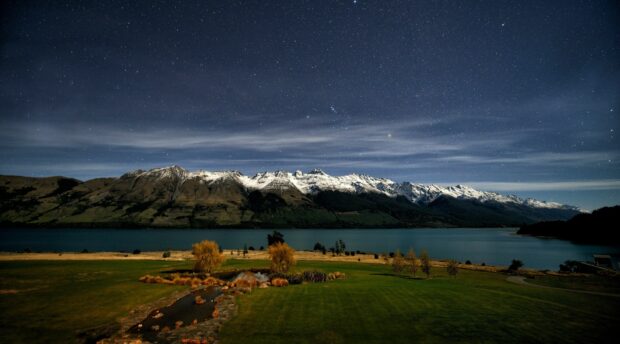 Snow capped mountains in New Zealand under a starry night sky with green fields and a lake in the foreground
