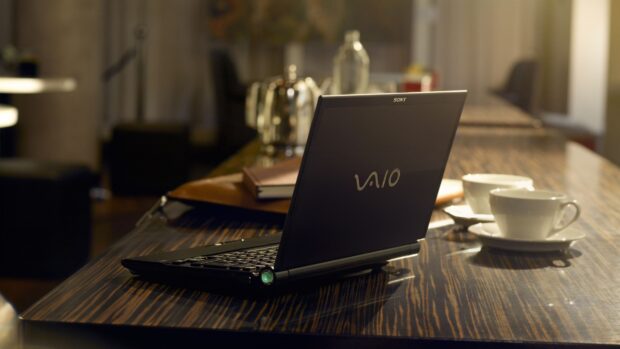 A black Sony VAIO notebook on a wooden table with cups and a book in the background