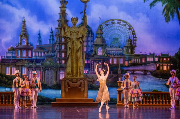 A ballet dancer performing on stage in a Nutcracker performance with a colorful cityscape backdrop