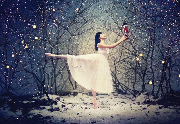 A ballet dancer holding a nutcracker on one leg in a snowy forest with sparkling lights