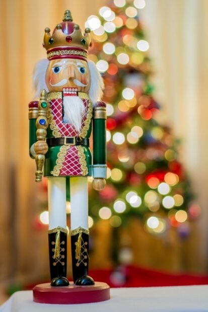 A detailed nutcracker figure standing in front of a decorated Christmas tree with festive lights