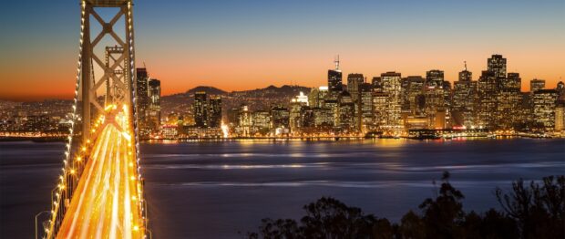 Evening cityscape featuring the Oakland skyline with illuminated bridge lights and sunset sky