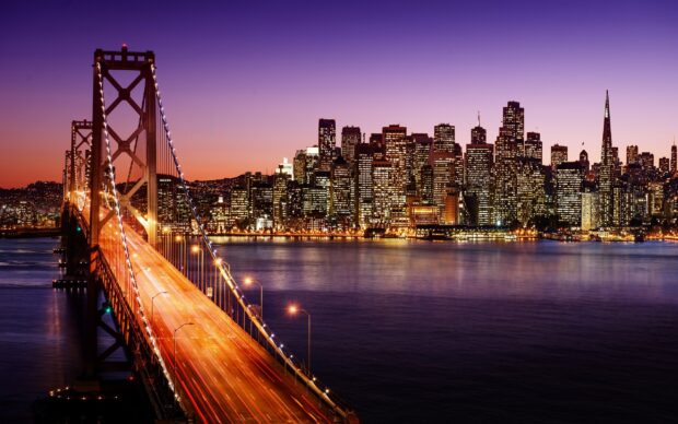 The Oakland skyline features illuminated buildings and a glowing bridge at dusk in the evening sky