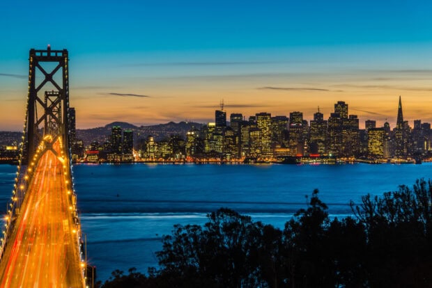 The Oakland skyline view at dusk with city lights glowing and a busy bridge in the foreground
