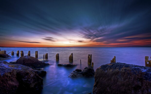 Calm ocean landscape with rocks and wooden posts at sunset sky displaying vibrant colors