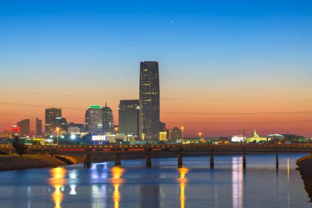 The Oklahoma City skyline features tall buildings and a bridge reflecting on the calm river at sunset