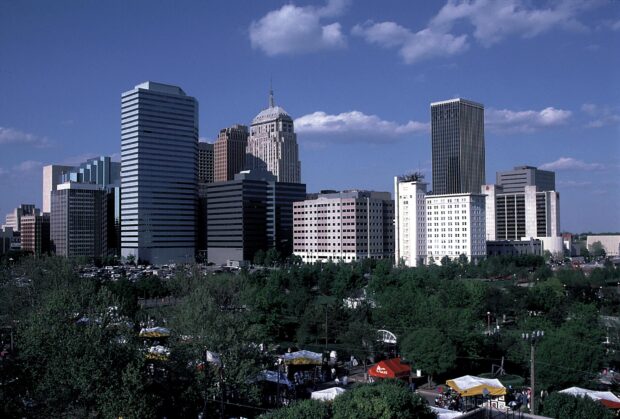 The Oklahoma City skyline features tall buildings under a clear blue sky with green trees in the foreground
