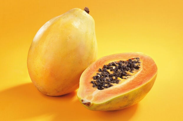 A fresh papaya fruit with one cut open showing seeds on a yellow background