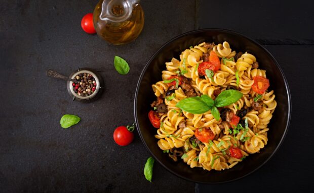 A plate of pasta with cherry tomatoes olives and fresh basil leaves on a dark surface