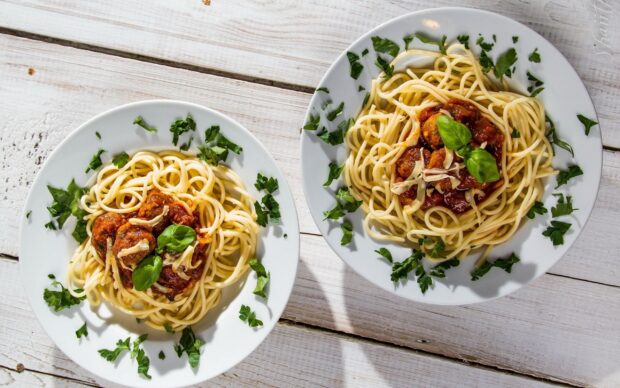 Two plates of spaghetti pasta with meatballs and fresh herbs served on a white wooden table