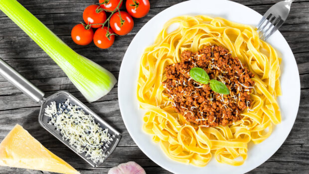 Fresh pasta with meat sauce and grated cheese on a plate with celery and tomatoes on a wooden table