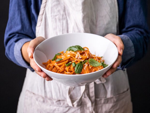 A person holding a bowl of freshly made pasta with basil leaves and grated cheese