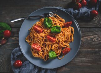 A plate of pasta with tomato and basil leaves on a wooden table with a fork and garlic cloves