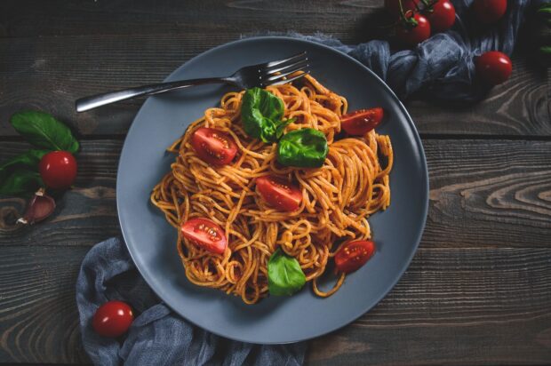 A plate of pasta with tomato and basil leaves on a wooden table with a fork and garlic cloves
