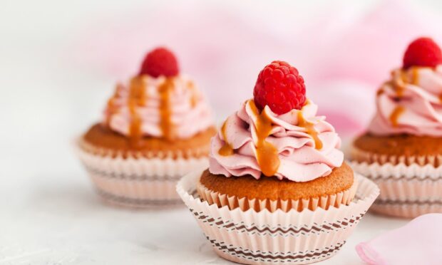 A raspberry topped pastry with pink cream and caramel drizzle in a decorative paper cup