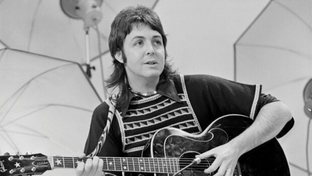 Paul Mccartney playing guitar in a studio setting wearing a patterned sweater