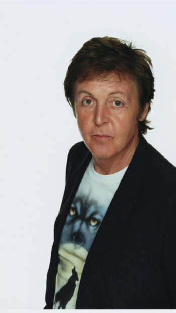 Paul Mccartney wearing a wolf printed shirt and black jacket standing against a white background