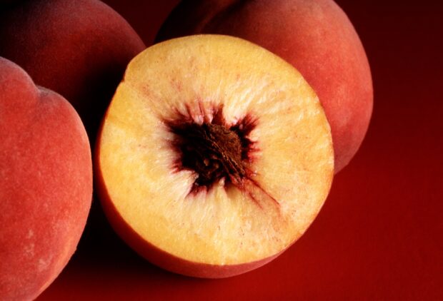 Fresh peach with seed cut in half surrounded by whole peaches on a red surface