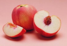 A fresh peach and sliced peach pieces showing the detailed fruit texture on a soft pink surface