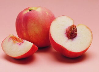 A fresh peach and sliced peach pieces showing the detailed fruit texture on a soft pink surface