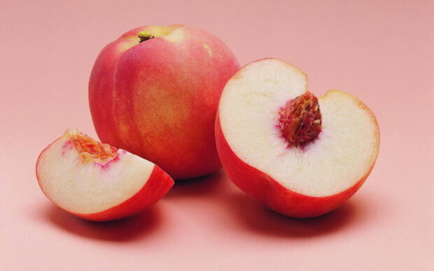 A fresh peach and sliced peach pieces showing the detailed fruit texture on a soft pink surface