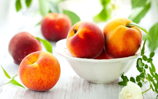 Fresh ripe peach fruit arranged in a white bowl on a wooden table surrounded by green leaves