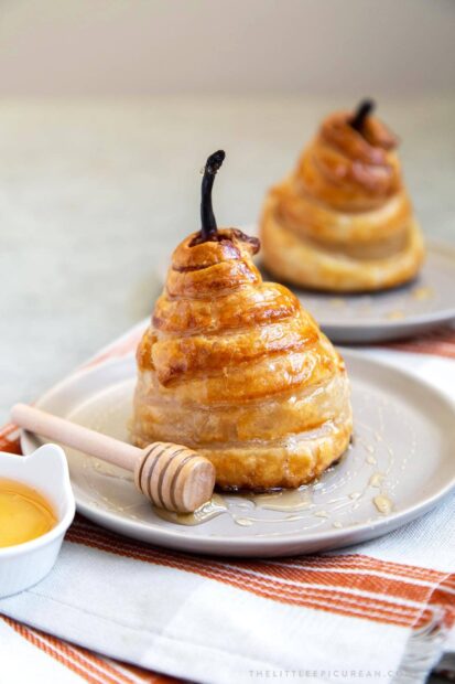 A golden baked pear wrapped in pastry with a honey dipper on a plate