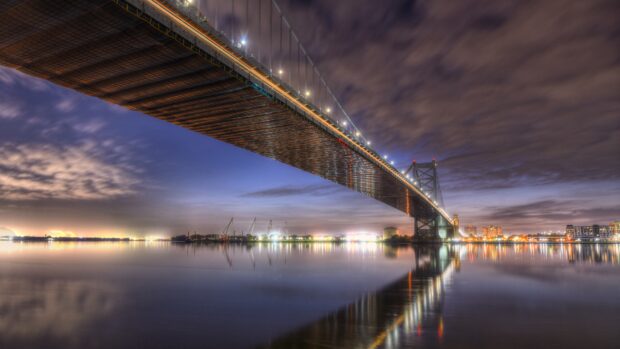 A large steel bridge in Pennsylvania spanning over calm water at dusk with city lights reflecting on the surface