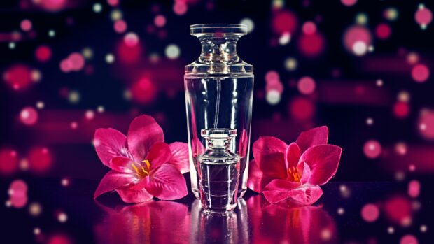 A clear glass perfume bottle surrounded by pink flowers on a reflective surface