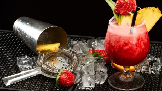A fresh pineapple slice next to a strawberry cocktail with ice and bar tools on the counter