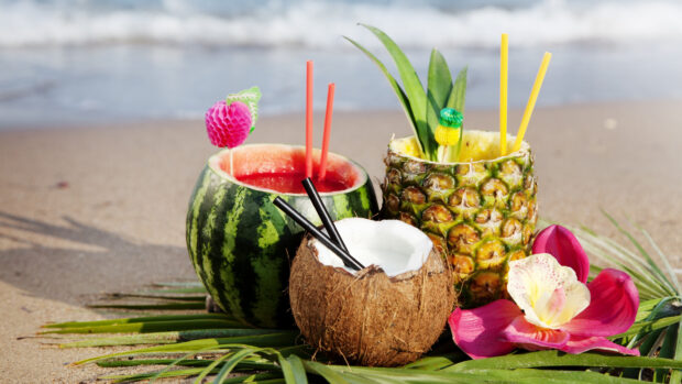 A pineapple filled with a tropical drink next to a coconut and watermelon on a beach