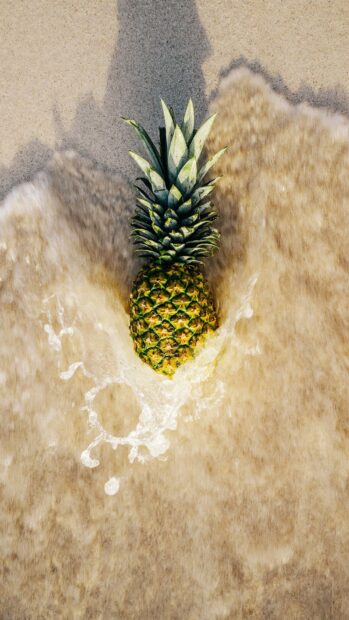 A ripe pineapple lying on wet sand with ocean water splashing around it