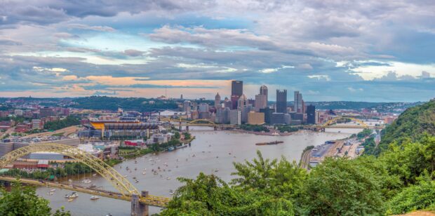 A panoramic view of Pittsburgh with bridges and river beside green hills at sunset