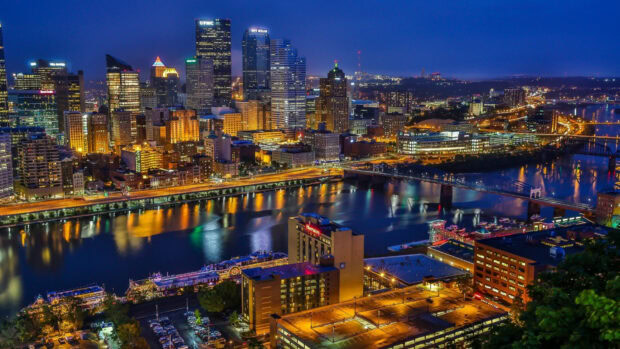 Nighttime cityscape of Pittsburgh skyline with illuminated buildings and river reflections