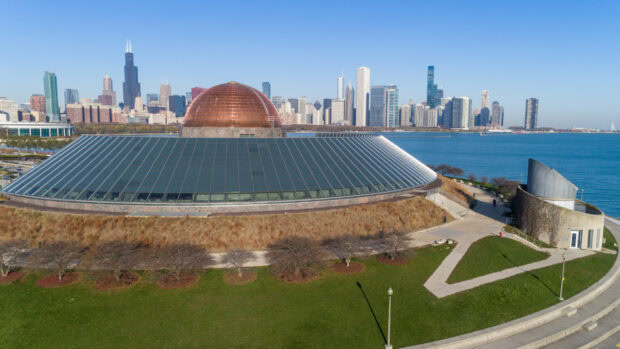 A copper dome planetarium surrounded by glass panels with a city skyline in the background