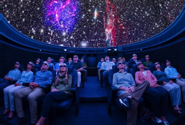 People wearing 3D glasses watching a planetarium projection ceiling filled with stars and colorful nebulae