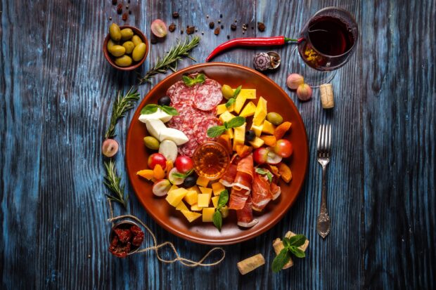 A plate filled with assorted meats cheese and olives arranged on a wooden table with herbs and a glass of red wine