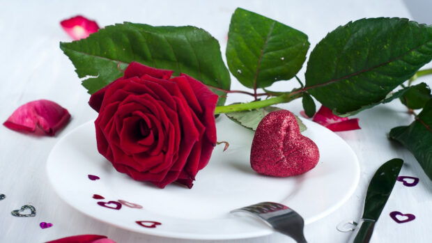 A red rose and a glittery heart placed on a white plate surrounded by green leaves and rose petals