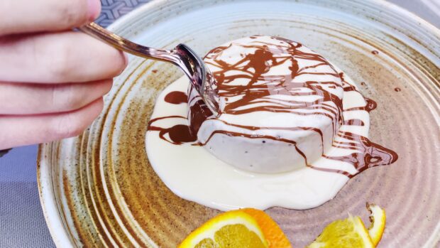 A hand holding a fork cutting a dessert on a plate with chocolate drizzle and orange slices