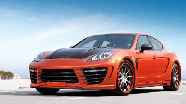 Orange Porsche Panamera with black hood parked on a beach parking lot under clear sky