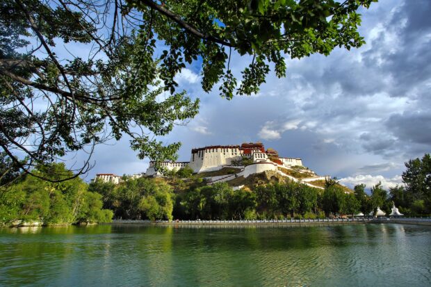 The Potala Palace surrounded by green trees and a calm lake under a cloudy sky