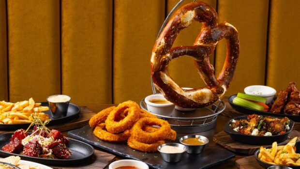 A large pretzel alongside fried onion rings and various appetizers on a wooden table