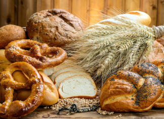 Fresh pretzel bread surrounded by various types of bread and wheat stalks on a wooden surface