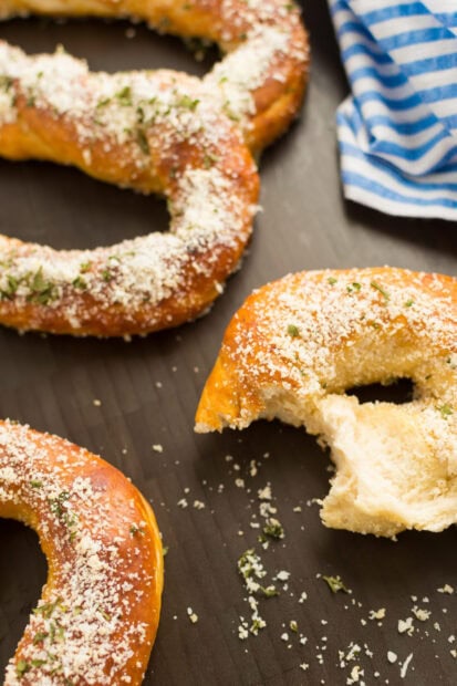 A close up of baked pretzel with cheese and herbs sprinkled on top on a dark surface