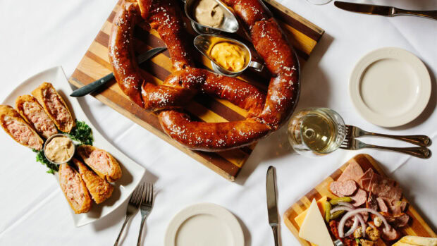 A large salted pretzel served on a wooden board with mustard dips and charcuterie sides