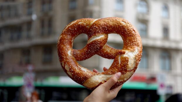A hand holding a salted pretzel in front of a blurred urban background